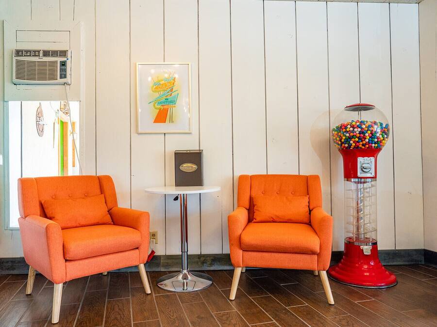 To orange lænestole, et rundt bord, et indrammet kunstværk, en rød gumball-automat og en hvid væg med lodrette paneler.