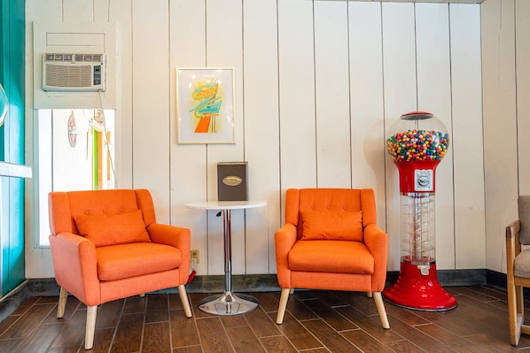 Two orange armchairs, a small round table, a framed artwork, a red gumball machine, and a white wall with vertical paneling.