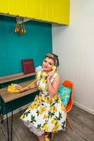 Uma pessoa sentada em uma cadeira em um quarto com parede verde-água, escrivaninha de madeira e telefone amarelo.