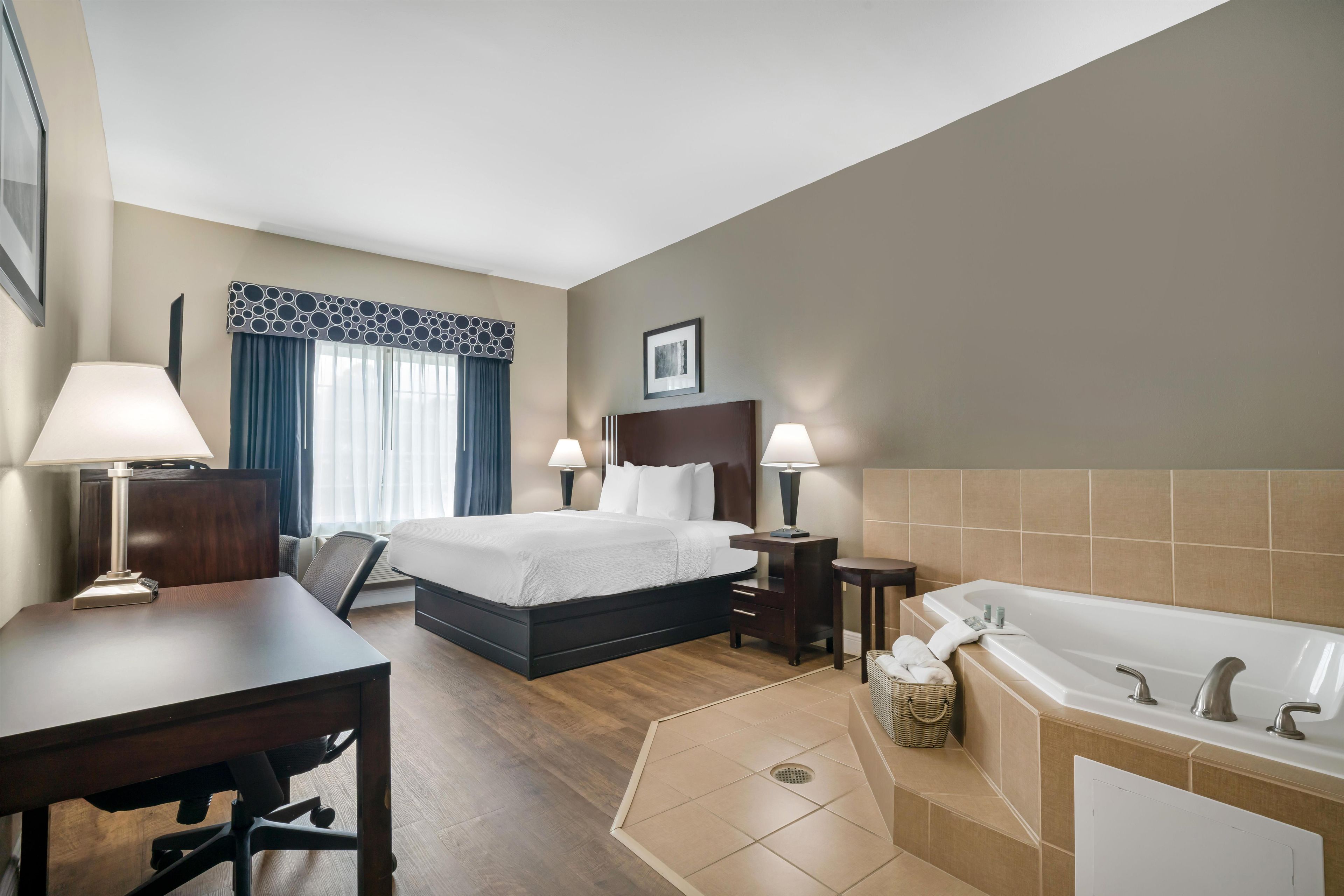 A hotel room with a bed, two bedside tables, a desk, a chair, a window with curtains, a framed picture on the wall, and a bathtub.