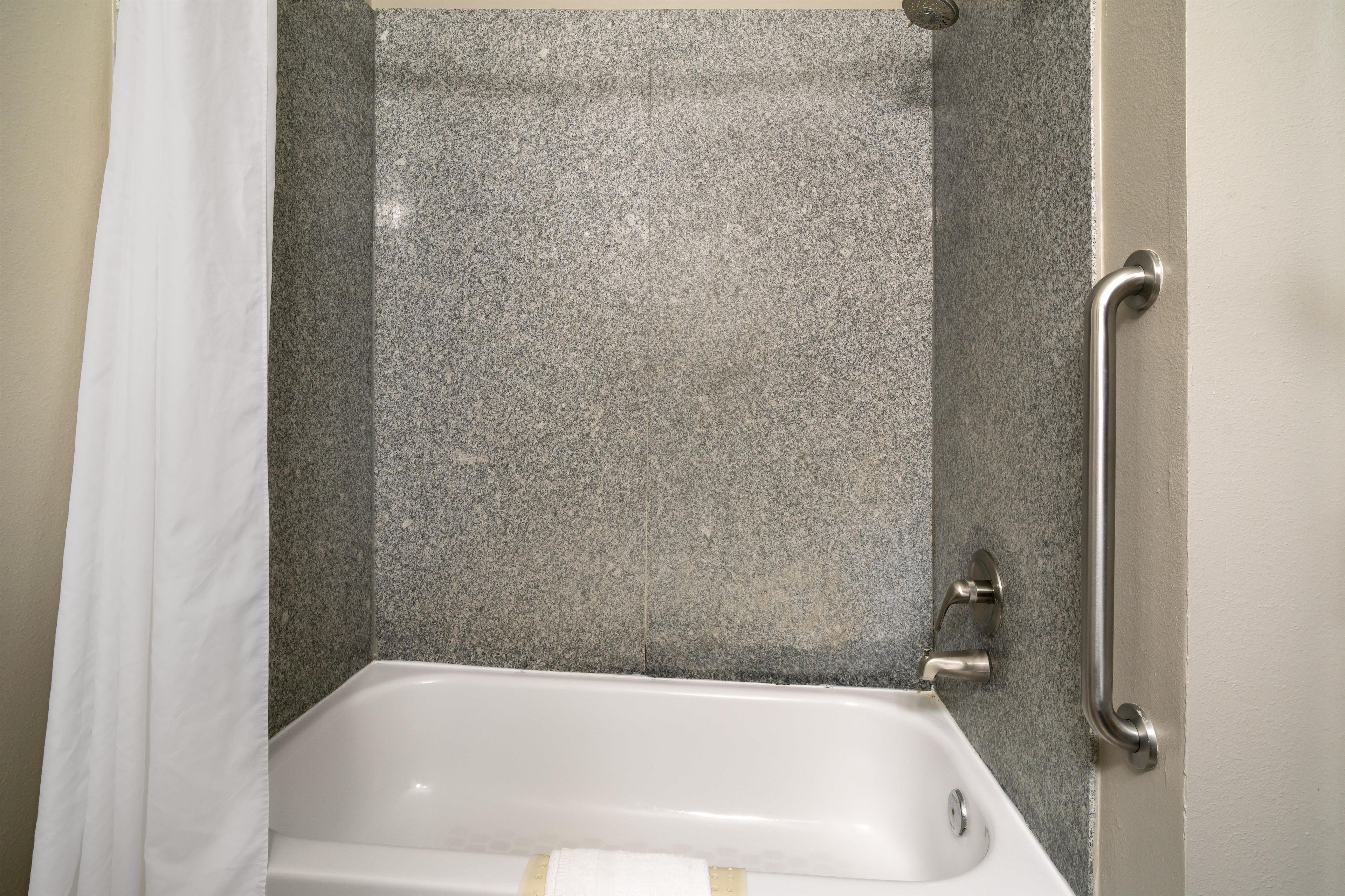 A bathroom with a white bathtub, a shower with a granite wall, and a grab bar on the right side.