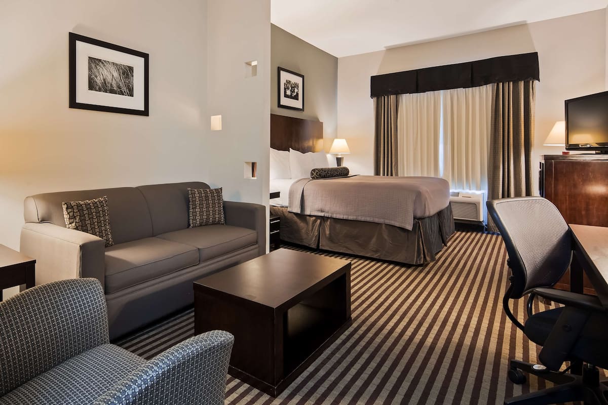 A hotel room with a sofa, two armchairs, a small table, a bed with a headboard, a desk, a lamp, and a picture frame on the wall.