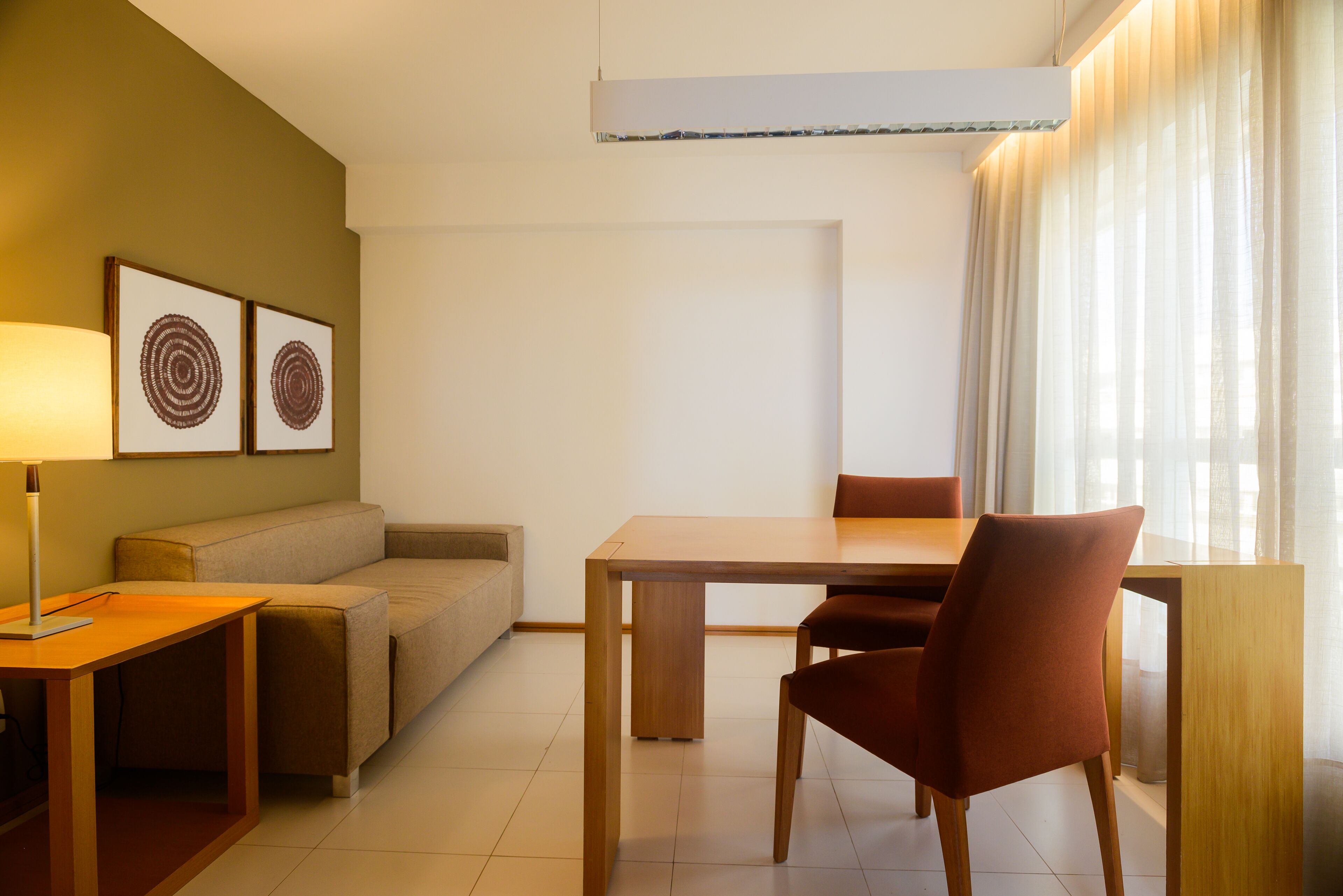Una habitación de hotel moderna con una mesa de comedor de madera, un sofá, una lámpara y dos cuadros en la pared.