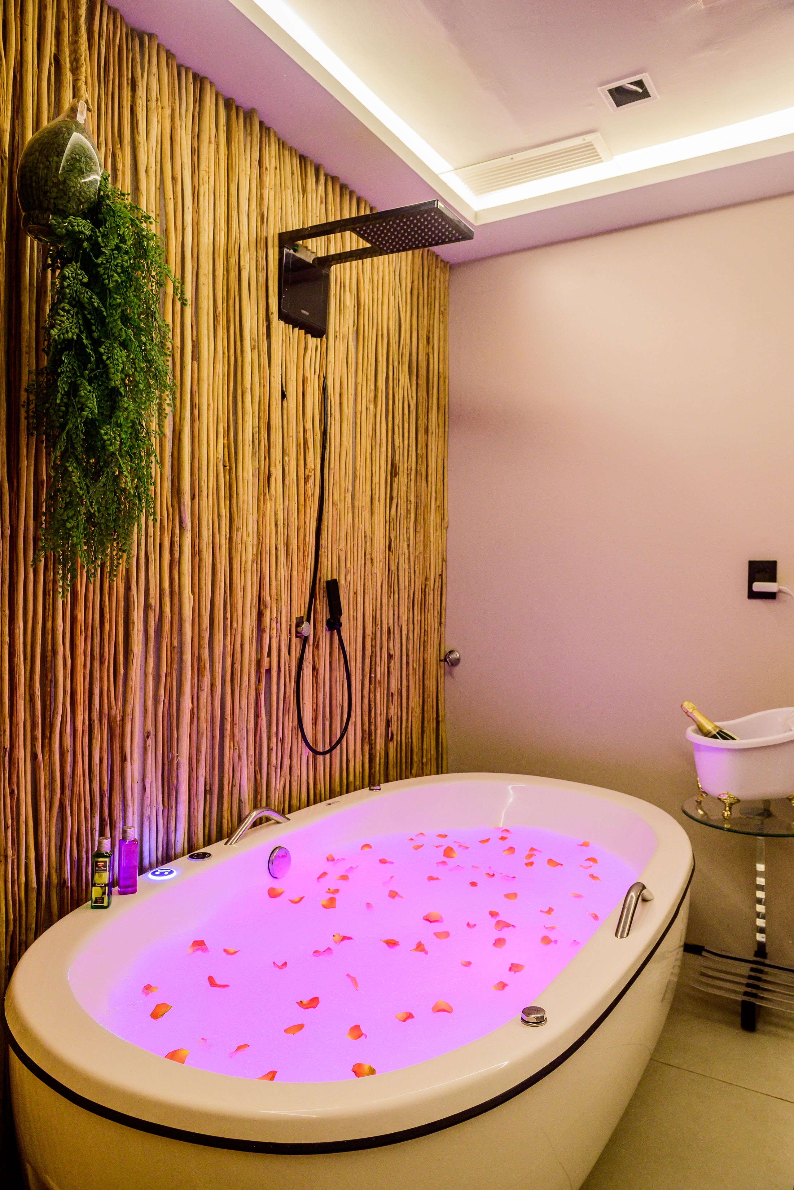 A large bathtub with rose petals, a wooden wall, and a small table with a bottle.