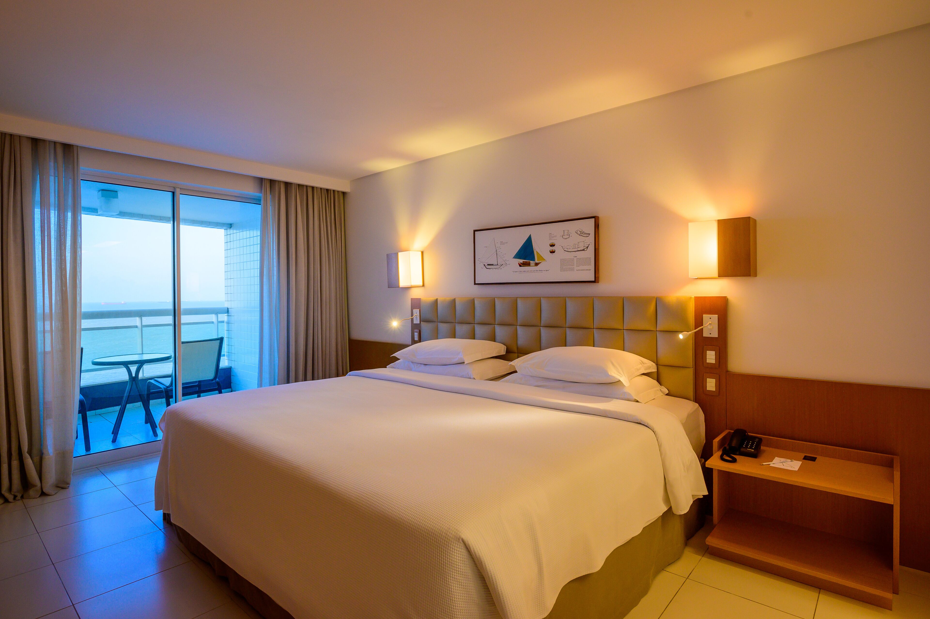A hotel room with a large bed, a bedside table, a chair, a window with curtains, and a view of the sea.