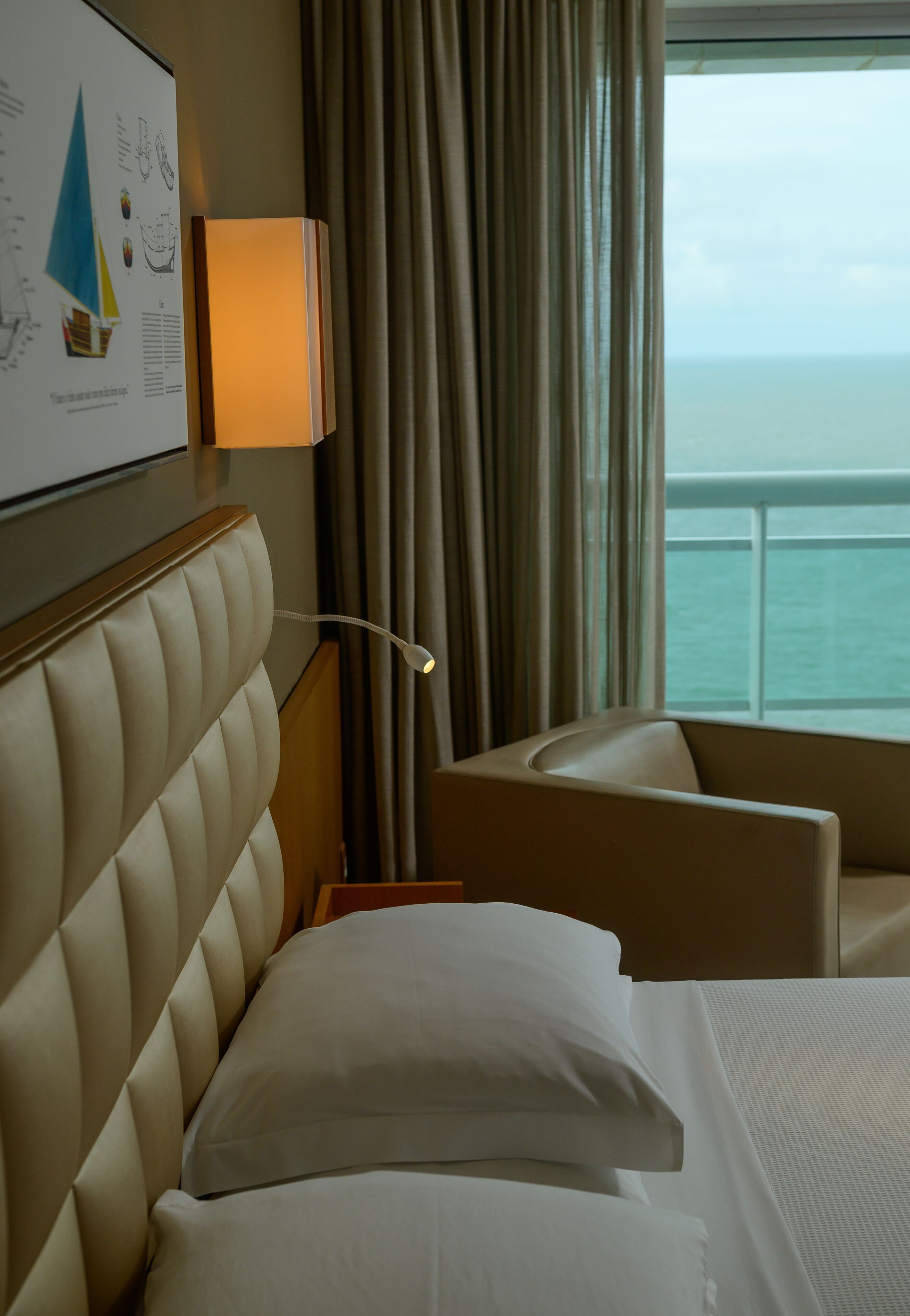 A hotel room with a bed, a chair, a wall-mounted lamp, and a window with curtains.