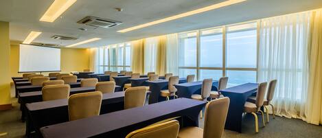 Un salón de conferencias con filas de mesas y sillas, ventanas amplias con cortinas y unidades de aire acondicionado en el techo.