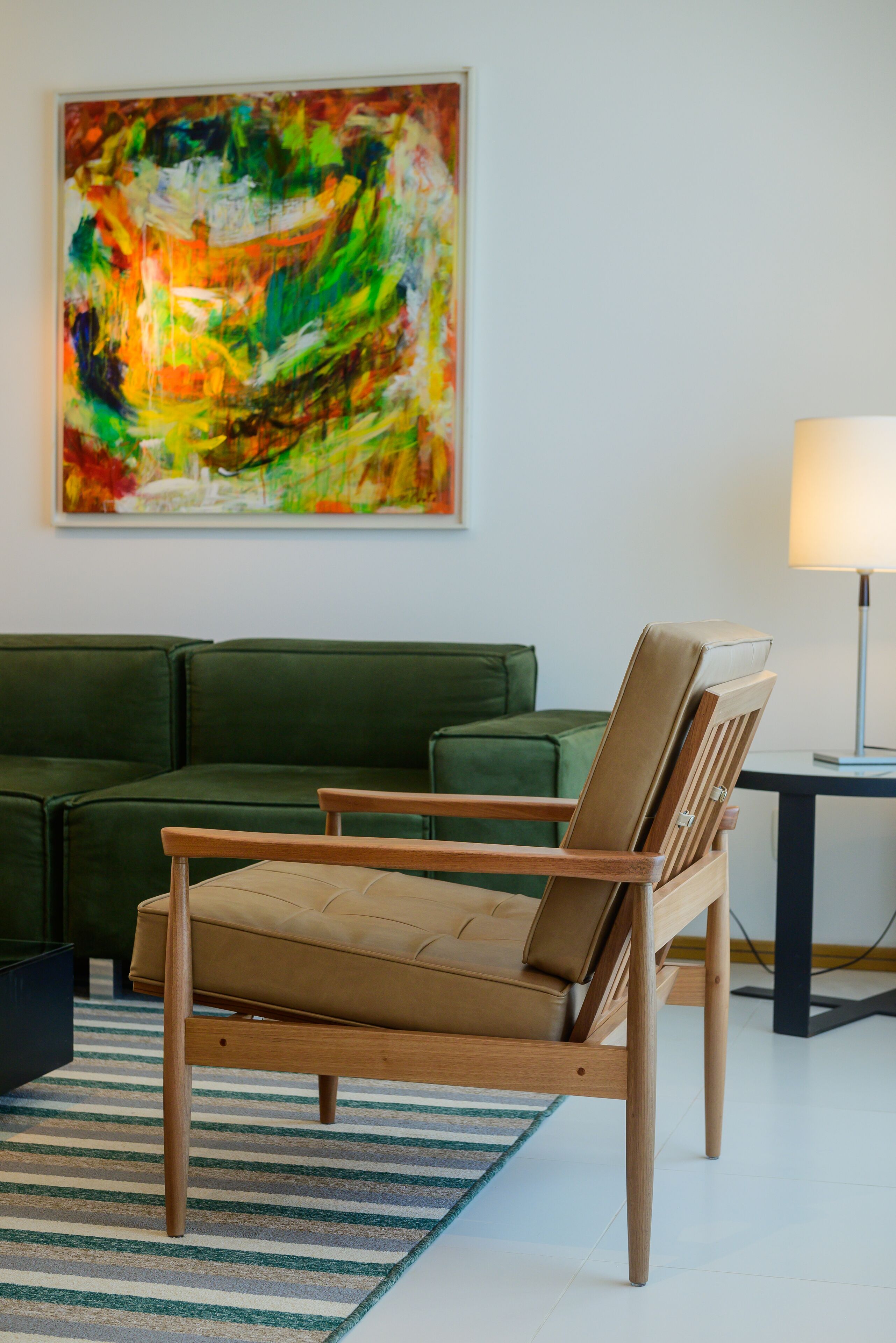 A room with a green sofa, a wooden chair, a lamp, and a colorful abstract painting on the wall.