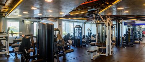 Un gimnasio con diferentes máquinas de ejercicio, amplias ventanas y un techo alto con conductos a la vista.