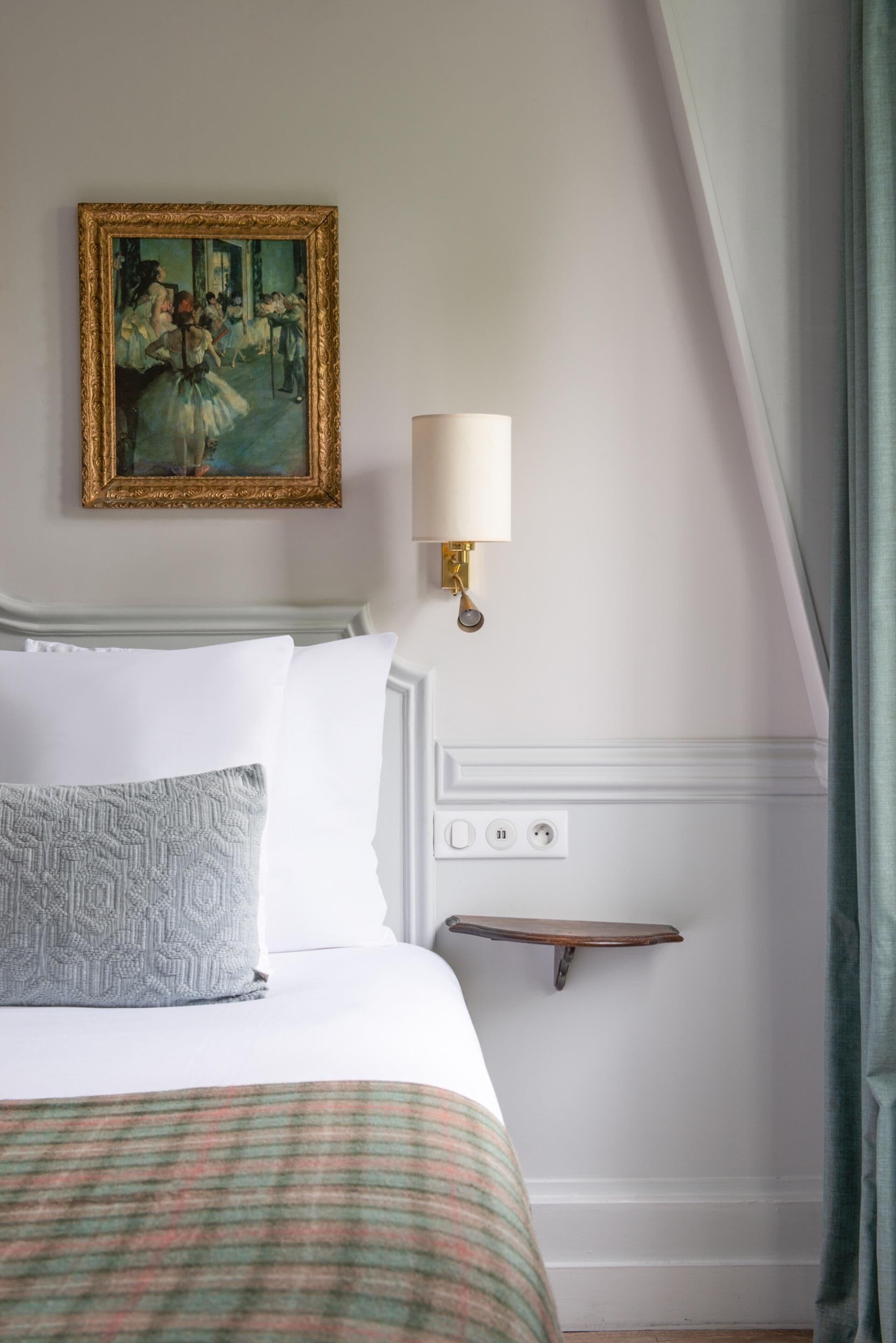 A hotel room with a bed, a painting, a wall-mounted lamp, and a power outlet.