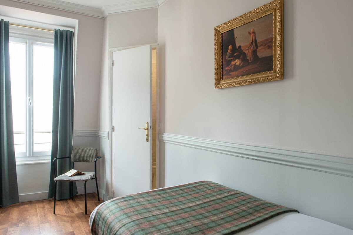 A room with a bed, a chair, a painting on the wall, and a window with curtains.