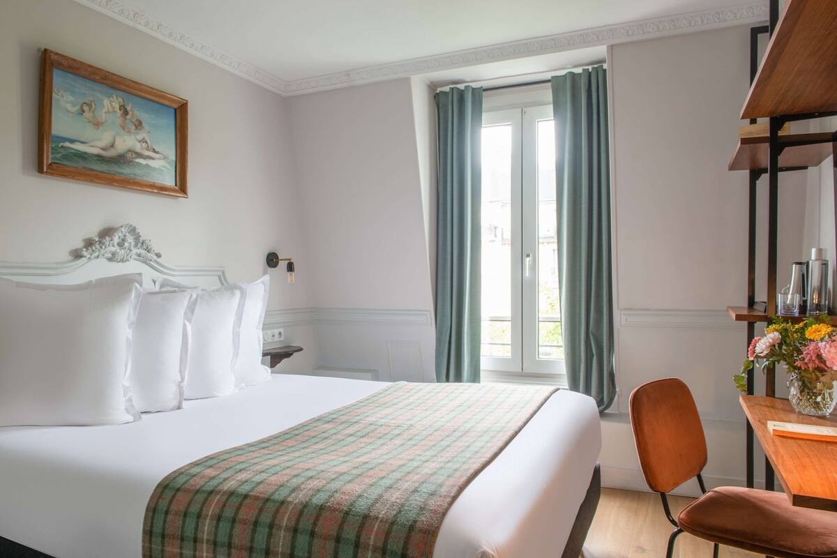 A hotel room with a bed, a desk, a chair, a window with curtains, and a painting on the wall.