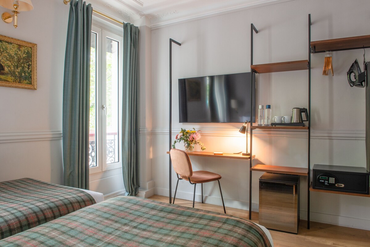 A hotel room with two beds, a desk with a chair, a flat-screen TV, a small kitchen area, and a window with curtains.