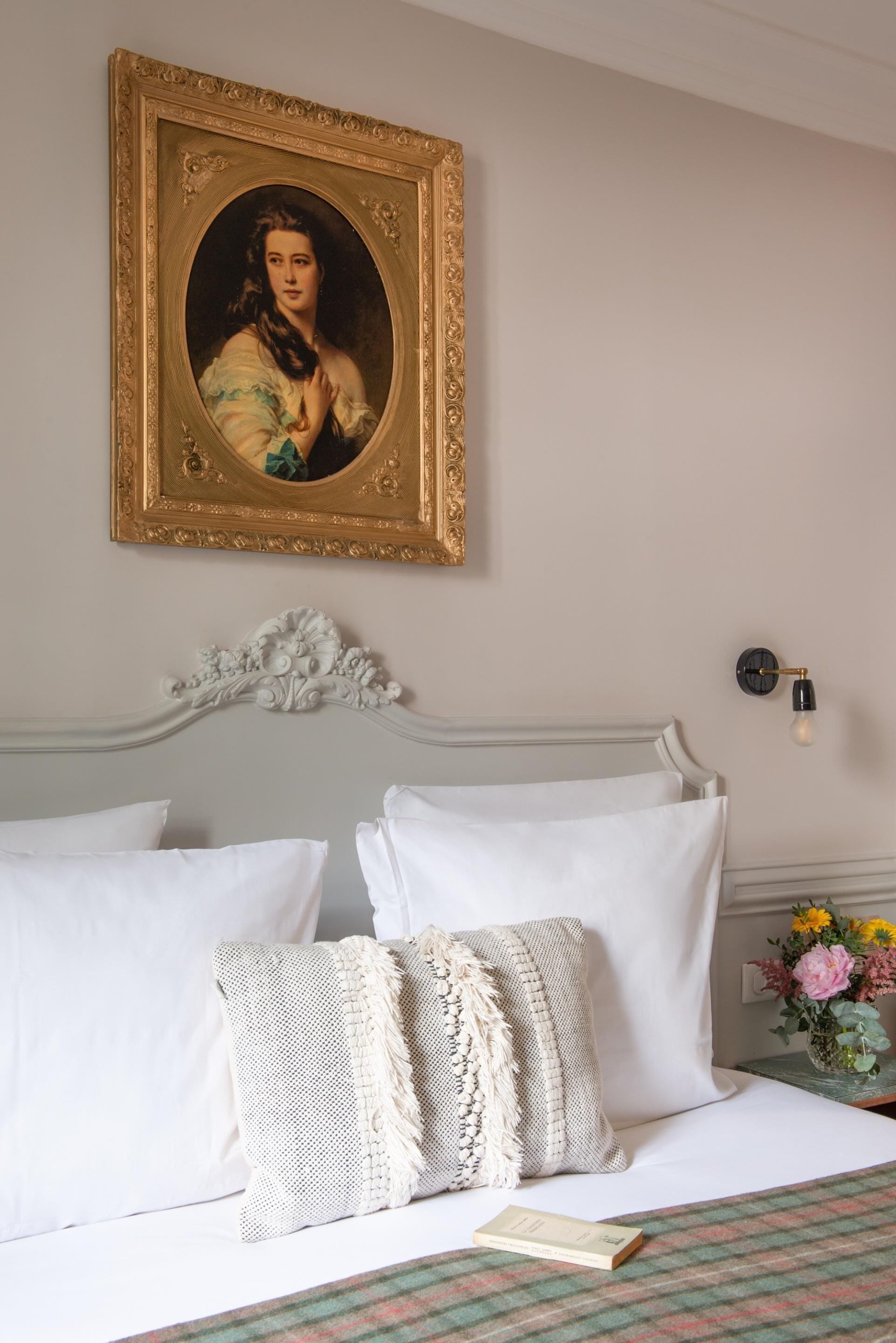 A bedroom with a bed, pillows, a framed portrait, a wall clock, and a vase with flowers.