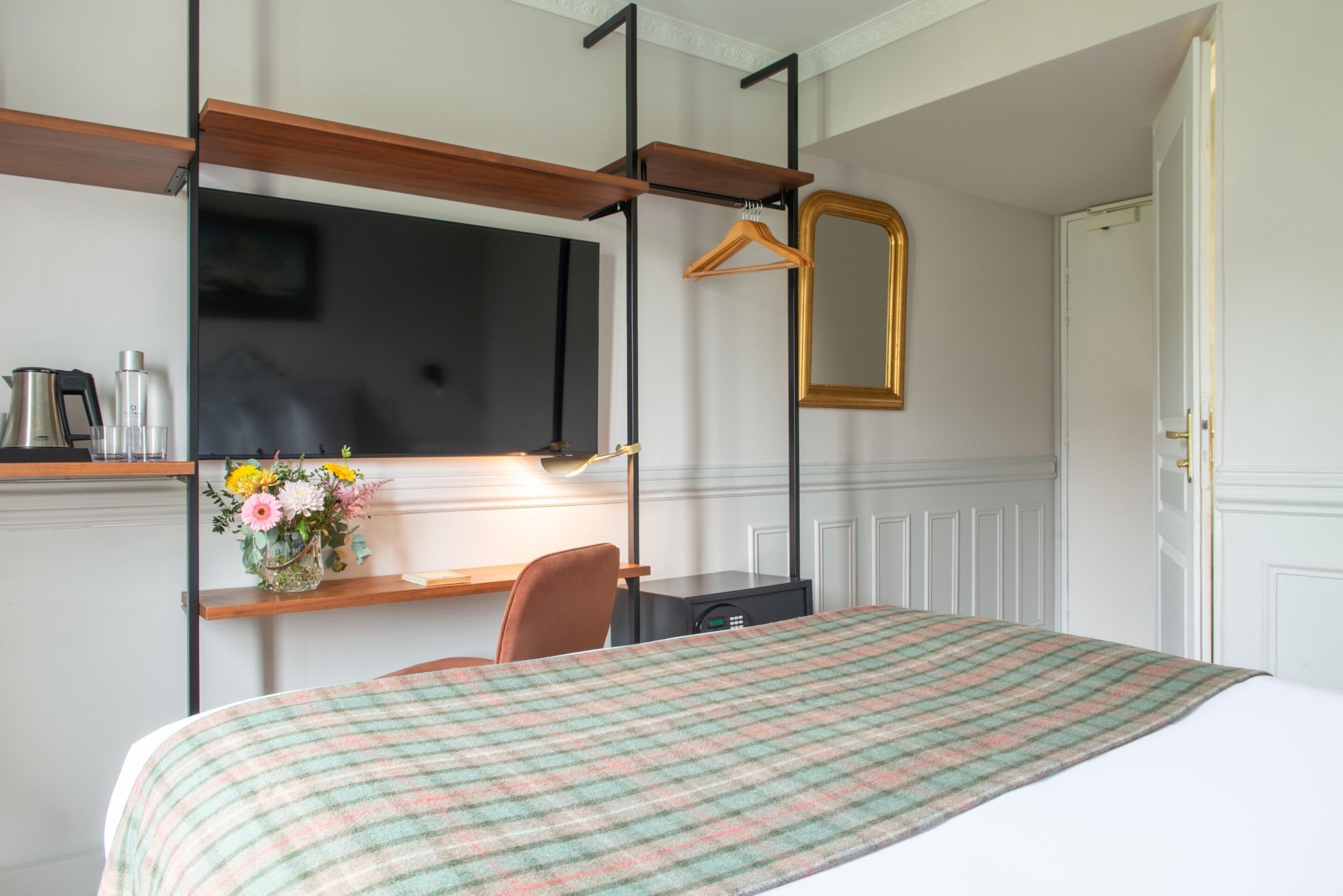 A hotel room with a bed, a desk with a chair, a TV mounted on the wall, and a mirror.