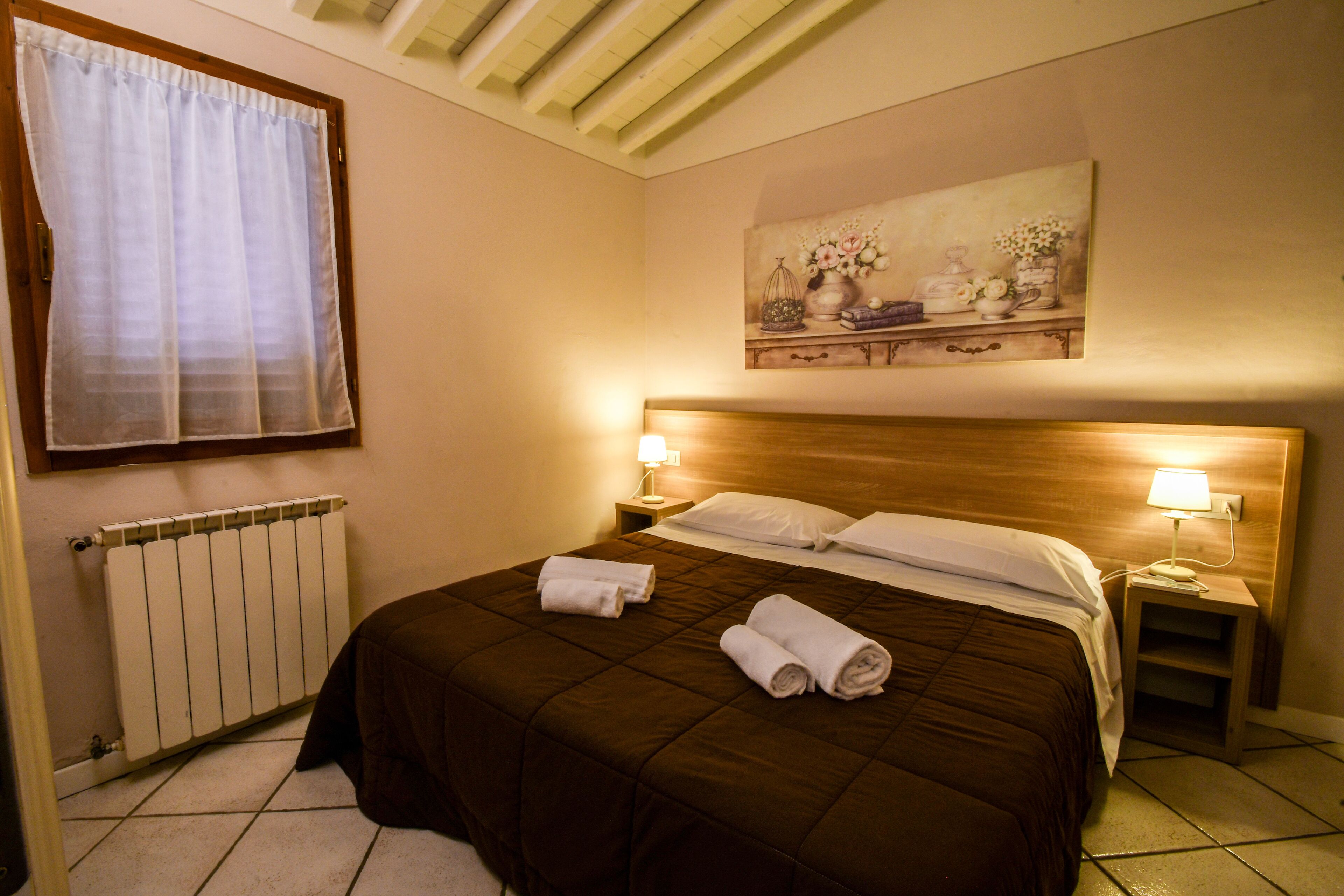 A hotel room with a large bed, two bedside lamps, a radiator, and a painting on the wall.