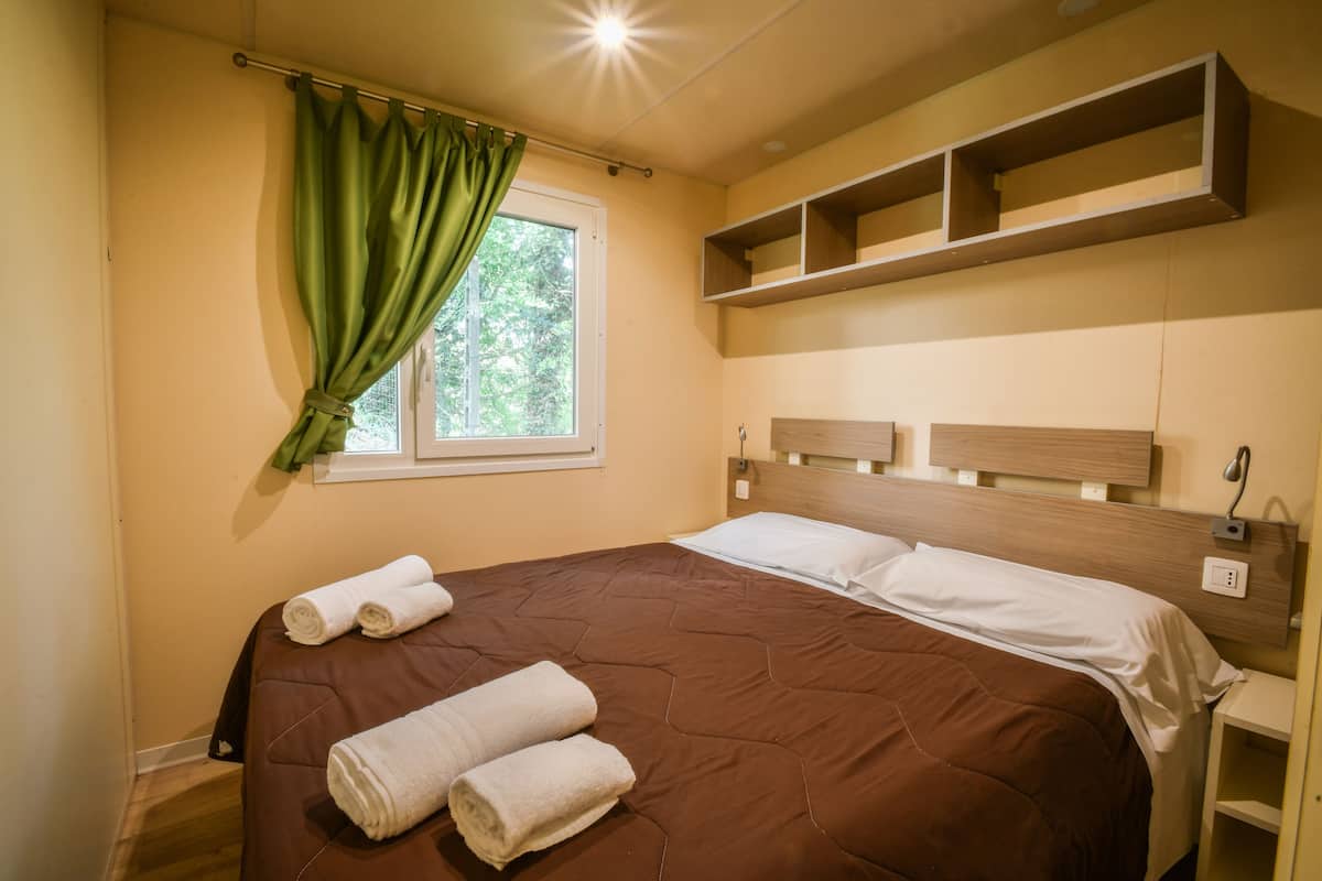 Un dormitorio con una colcha marrón, dos toallas enrolladas, una ventana con cortinas verdes y estantes de madera sobre la cama.