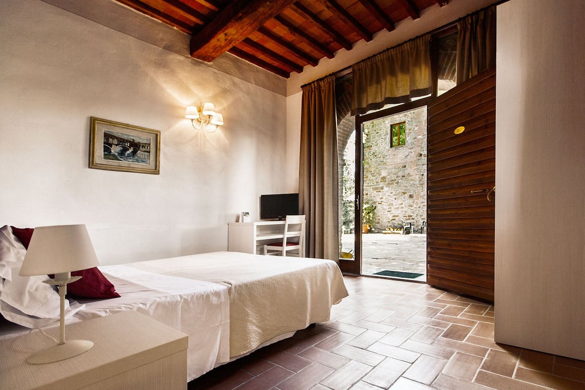 Un dormitorio con cama, escritorio, silla, televisor y vista a un área exterior con pared de piedra y puerta de madera.