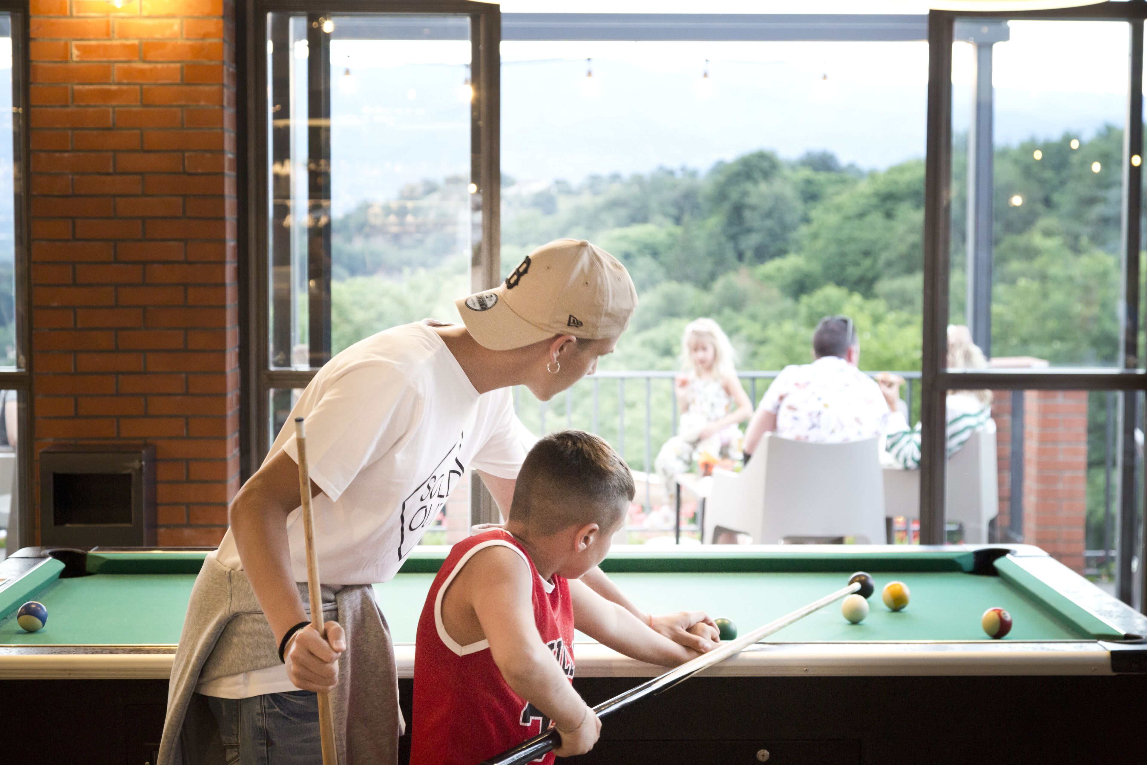 A pool table with balls, a person standing beside it, and a view of greenery through large windows.