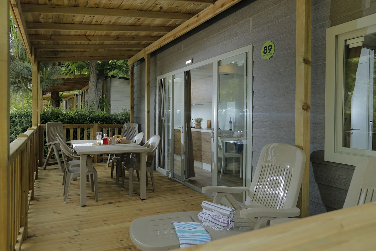 Un patio cubierto al aire libre con una mesa y sillas de madera, una silla reclinable y una puerta corrediza de vidrio que da a una zona de cocina.