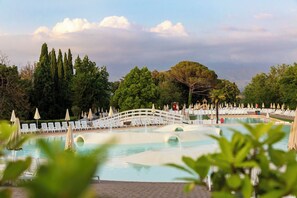 Zona de piscina con sillas de descanso y sombrillas, rodeada de árboles y un puente.