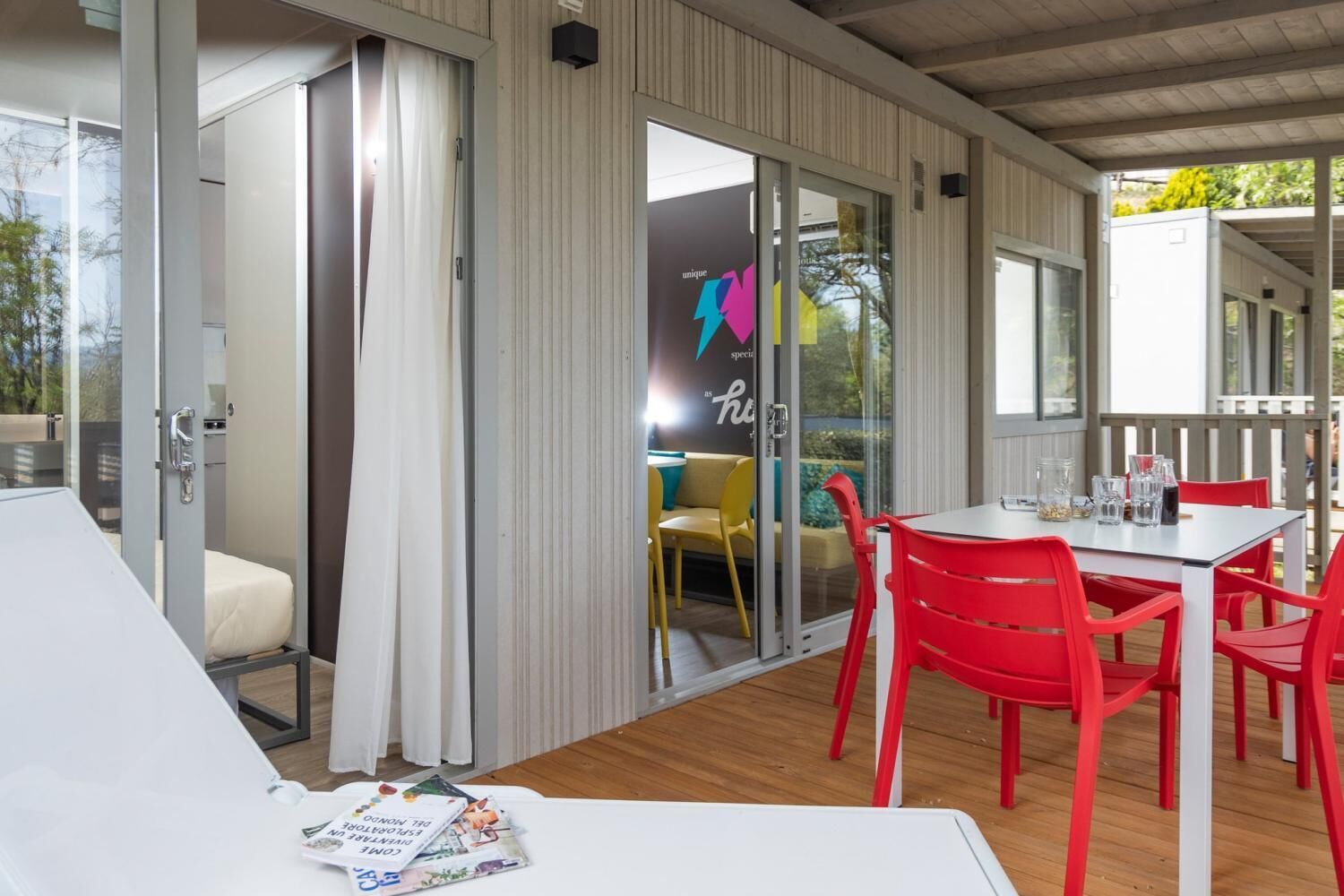 Un área de estar moderna al aire libre con una mesa blanca y sillas rojas, una puerta de vidrio que da a una silla amarilla, y vistas al exterior.