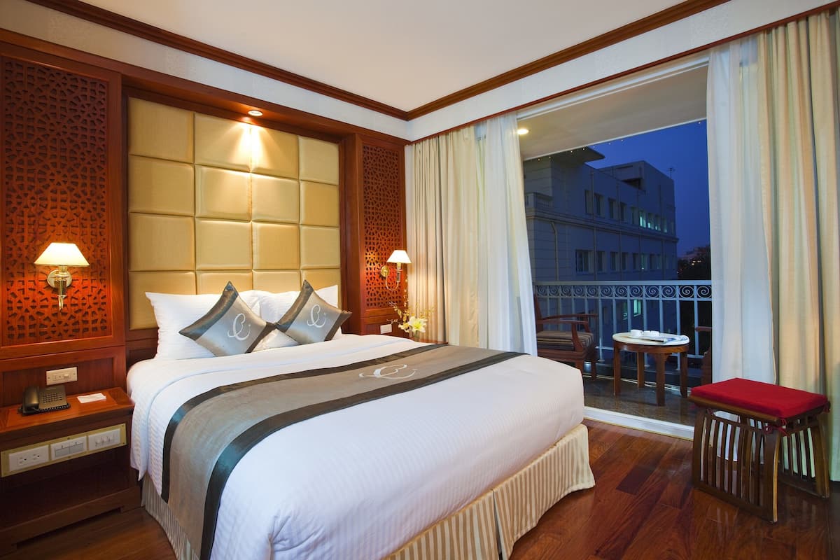 Habitación de hotel con una cama grande, cabecero de madera, balcón con vista a un edificio y una pequeña mesa con un cojín rojo.