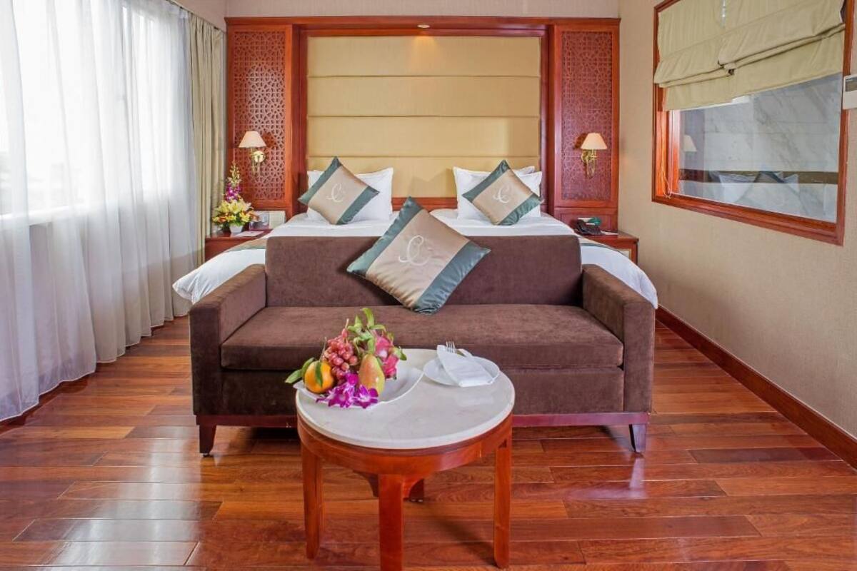 Una habitación de hotel con una cama grande, un sofá marrón, una mesa redonda de madera con una cesta de frutas y una ventana con cortinas.