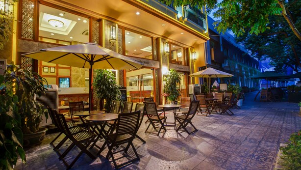An outdoor patio with tables and chairs, surrounded by potted plants and a building with large windows and decorative lighting. - Conifer Boutique Hotel (Hanoi)