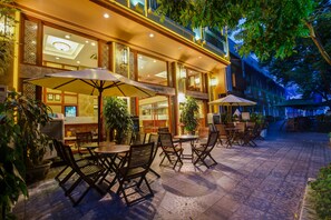 An outdoor patio with tables and chairs, surrounded by potted plants and a building with large windows and decorative lighting. - Conifer Boutique Hotel (Hanoi)