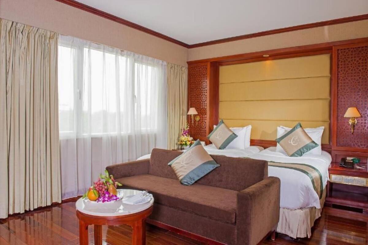 Una habitación de hotel con una cama grande, un sofá marrón, una mesita redonda con frutero y una ventana con cortinas.