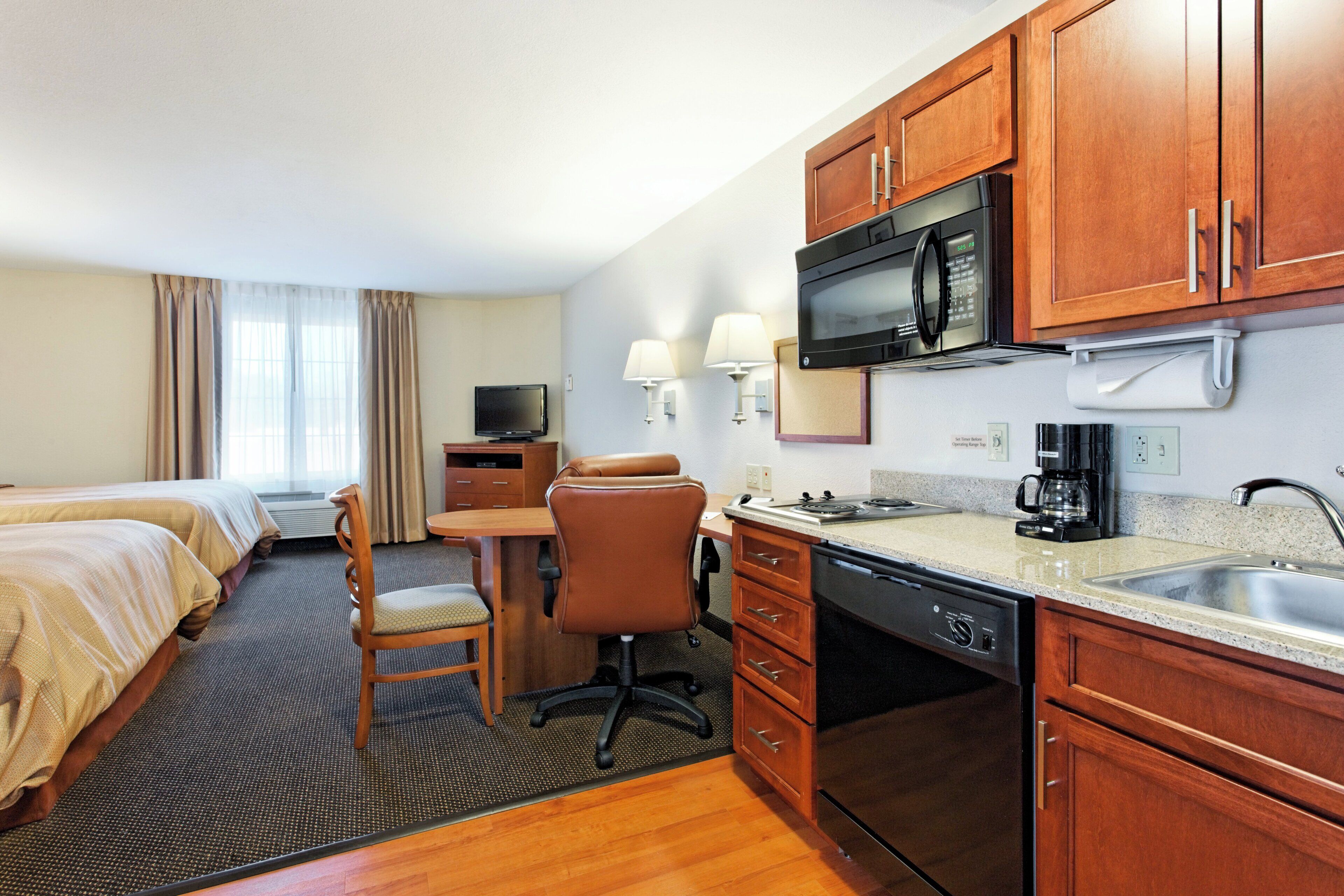 A hotel room with a kitchenette, a bed, a desk, a chair, and a television.