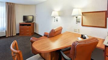 A hotel room with a wooden dining table, four leather chairs, a television on a wooden cabinet, two wall-mounted lamps, and a telephone.