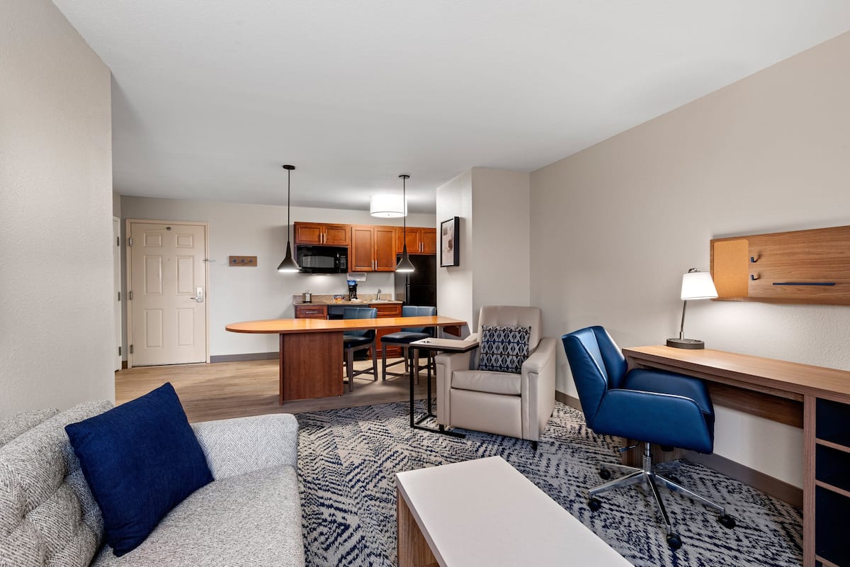 A modern living room with a sofa, a coffee table, a blue armchair, and a desk with a lamp. A kitchen area with wooden cabinets and a microwave is visible in the background.