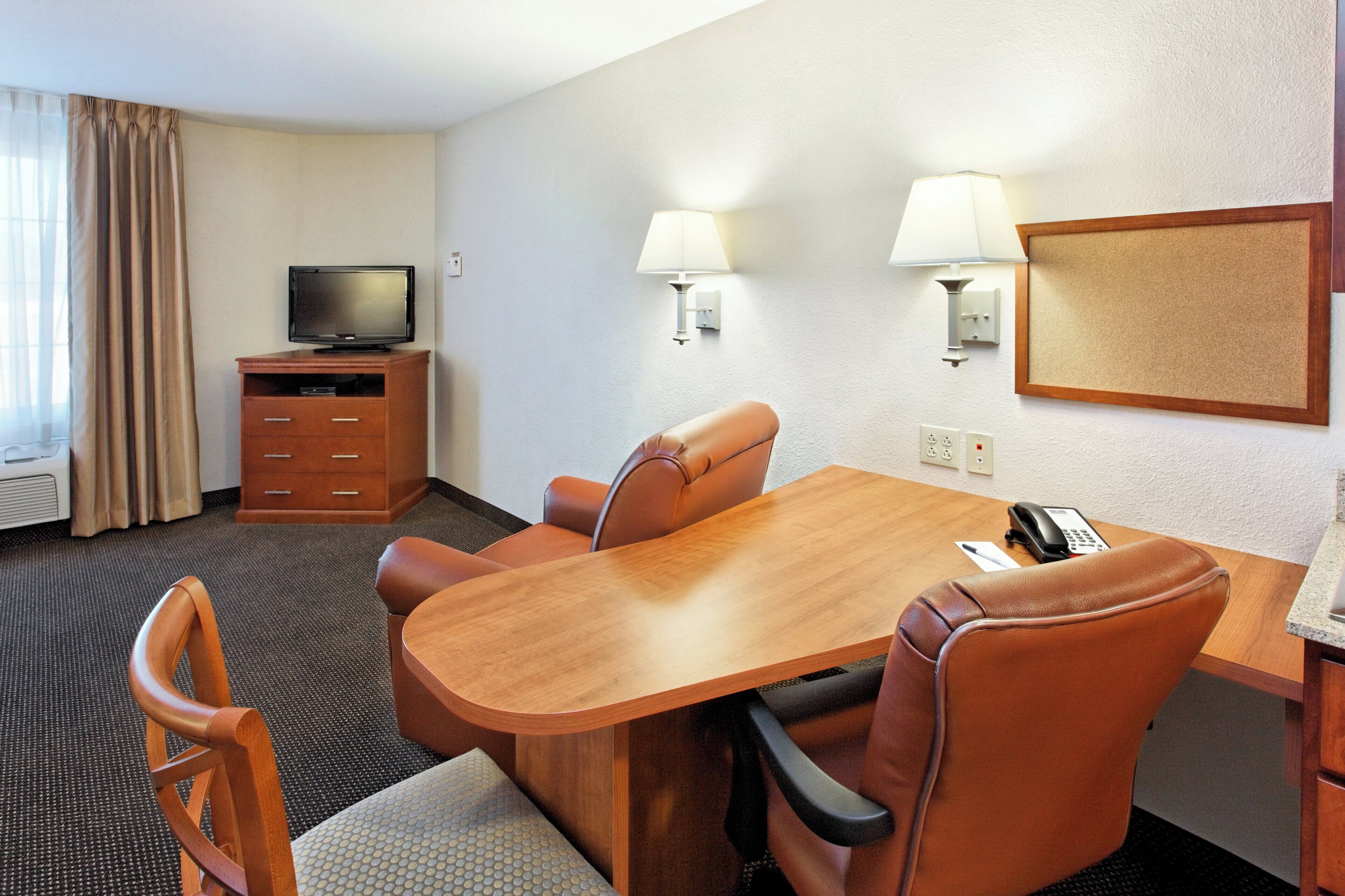 Une chambre d’hôtel avec un bureau en bois, deux chaises, un téléviseur posé sur un meuble, une lampe et un tableau d’affichage en liège accroché au mur.