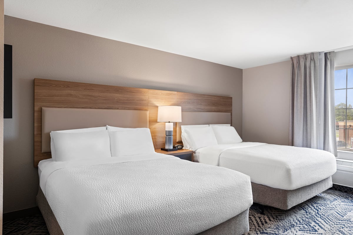 A hotel room with two beds, a wooden headboard, a nightstand with a lamp, and a view of the outdoors through a window.