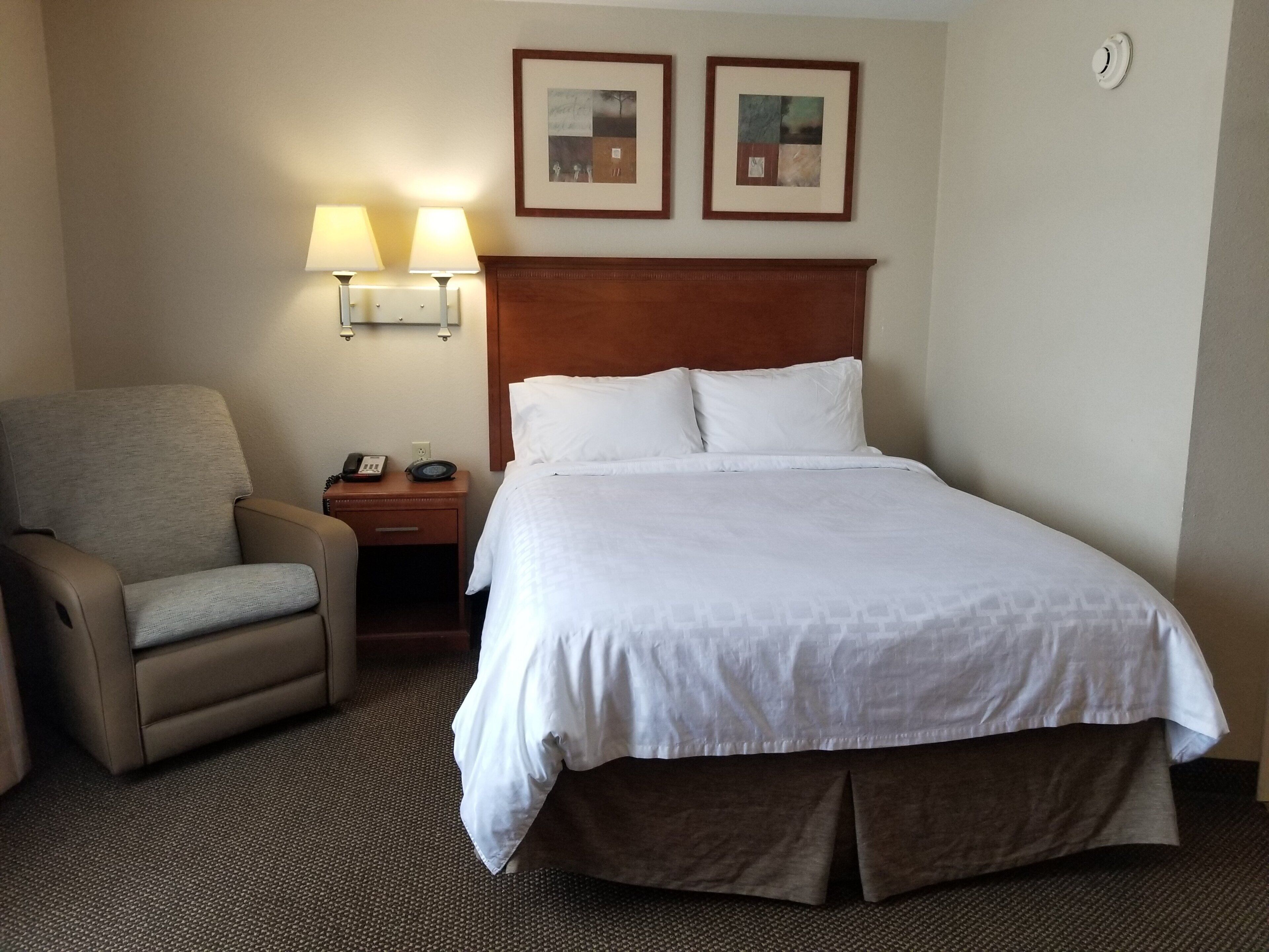 Une chambre d’hôtel avec un lit, une chaise, une table de chevet avec une lampe et deux tableaux encadrés au mur.