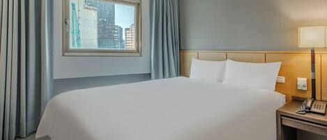 A hotel room with a large bed, a bedside table with a lamp, a window with curtains, and a view of buildings outside.