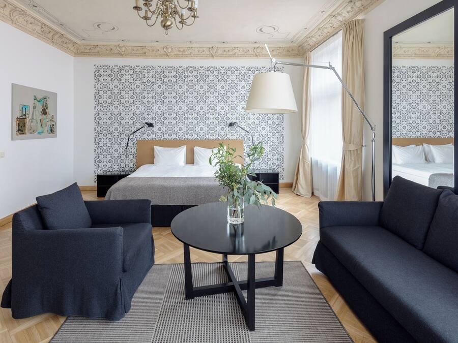 Una habitación de hotel moderna con una cama grande, dos sofás oscuros, una mesa redonda con un florero y papel tapiz estampado.
