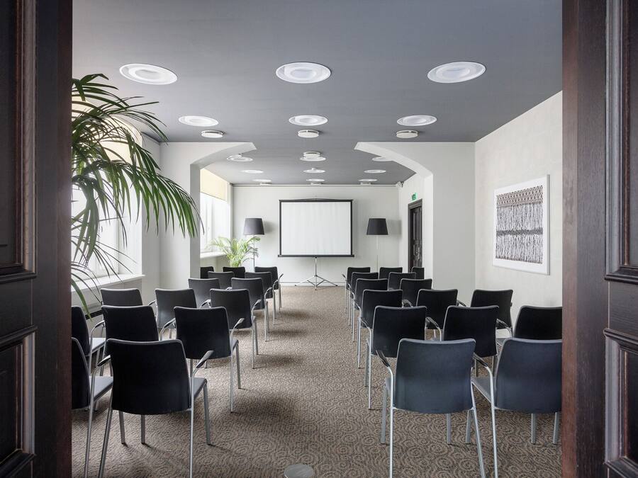 Un salón de conferencias con filas de sillas, una pantalla para proyector y una planta grande.