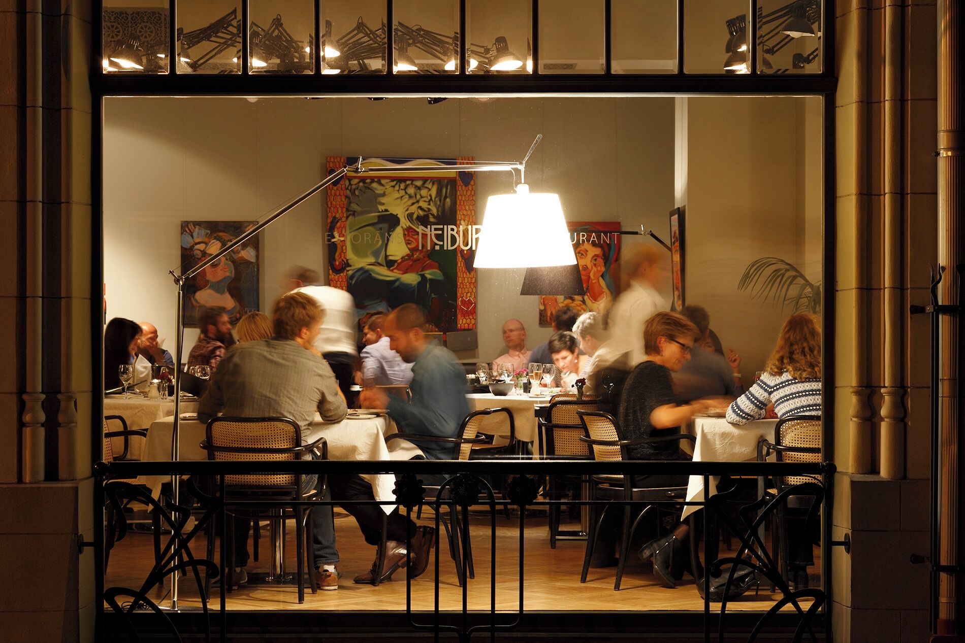Un restaurante con comensales dentro, que cuenta con un gran ventanal, una lámpara colgante y un mural en la pared.