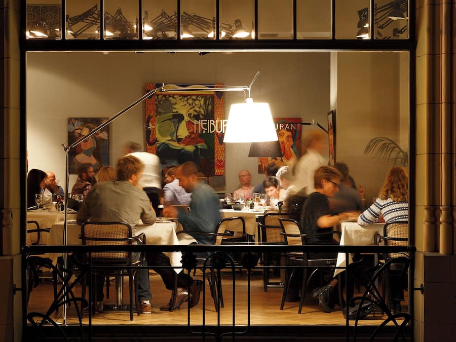 Un ristorante con avventori che cenano all'interno, caratterizzato da un'ampia vetrata, un lampadario e un murale sulla parete.