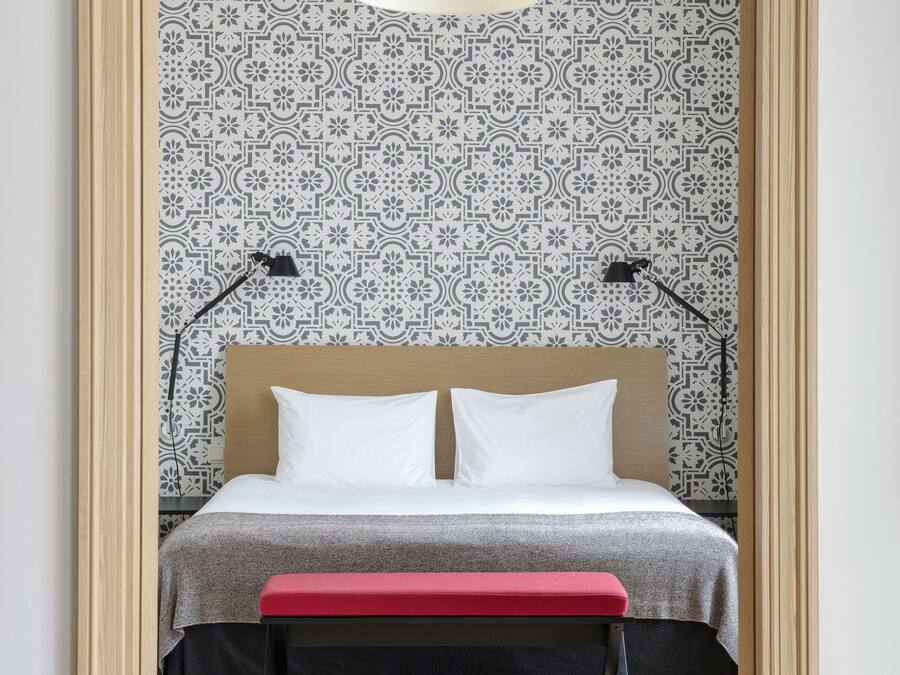A bedroom with a bed, a red bench, a hanging light, and patterned wallpaper.