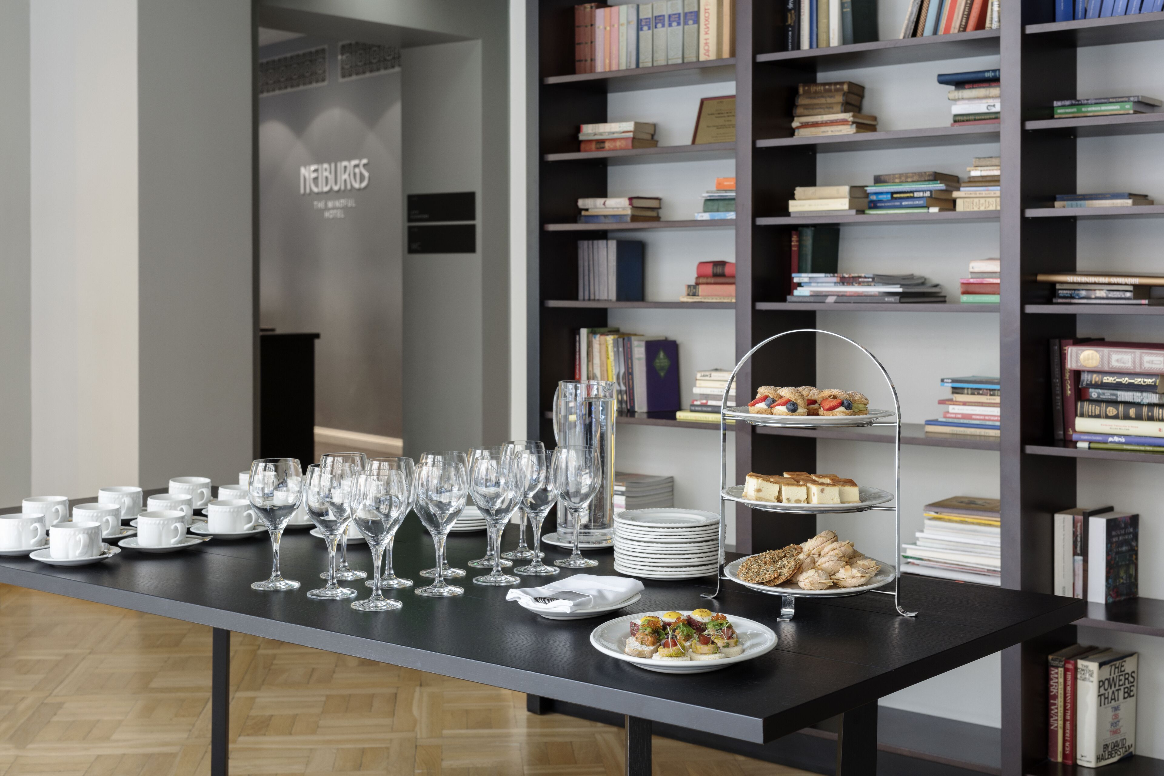 A table set with white cups, wine glasses, and plates of food, with a bookshelf in the background.