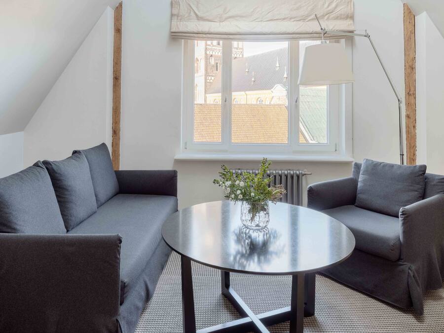 Una sala de estar moderna con dos sofás grises, una mesa redonda con un florero y una ventana con vistas a edificios.