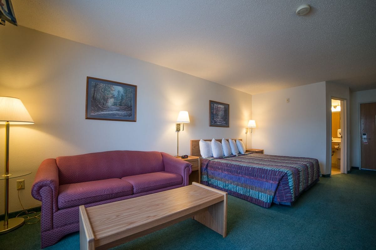 A hotel room with a purple sofa, a wooden bench, a bed with a colorful blanket, two bedside lamps, and a painting on the wall.