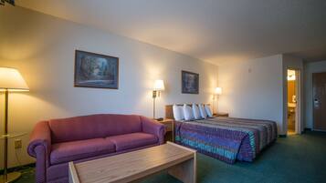 A hotel room with a purple sofa, a wooden bench, a bed with a colorful blanket, two bedside lamps, and a painting on the wall.