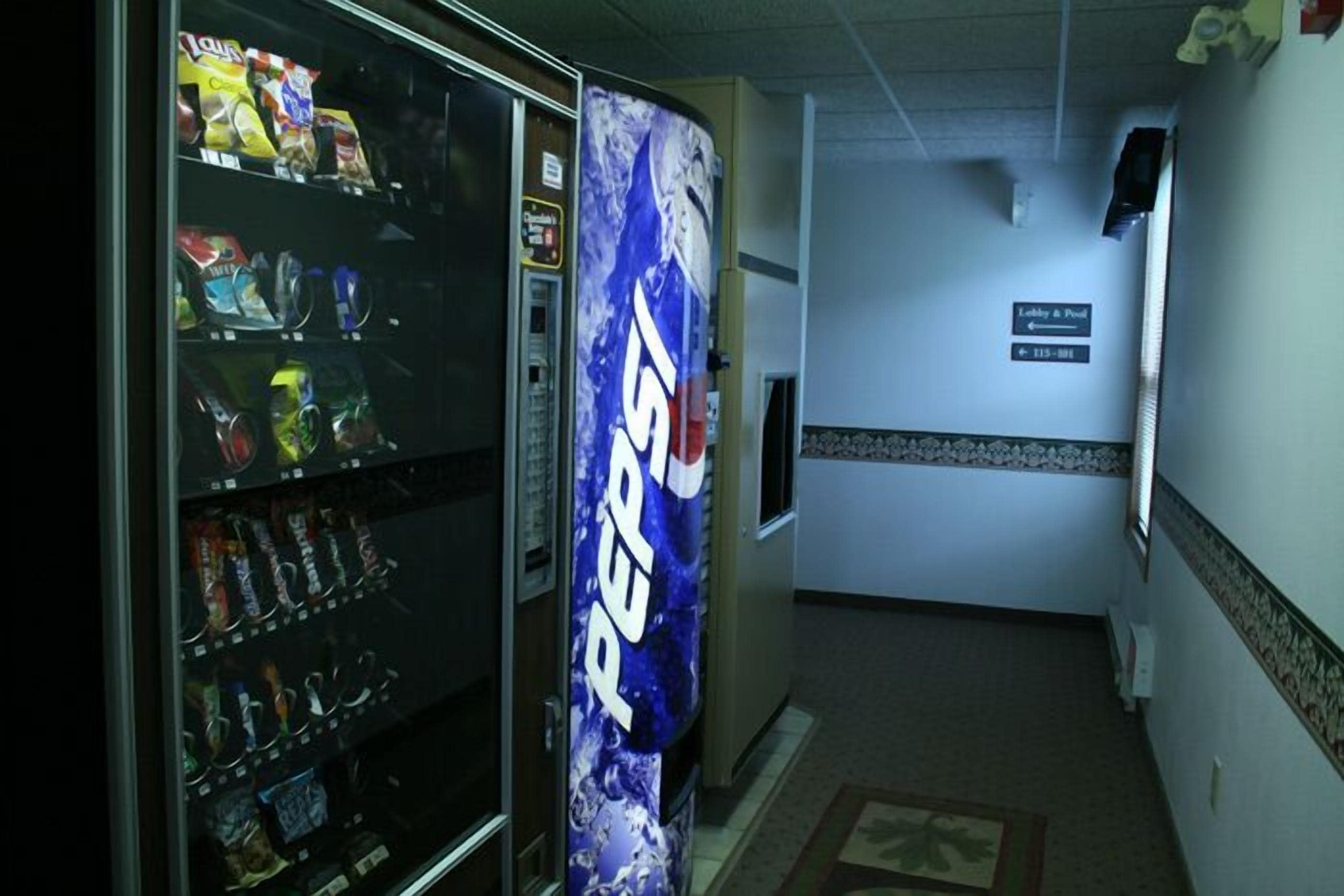 Un corridoio con una macchina distributrice piena di snack e una pubblicità di Pepsi. Un cartello indica la direzione verso la hall e la piscina.