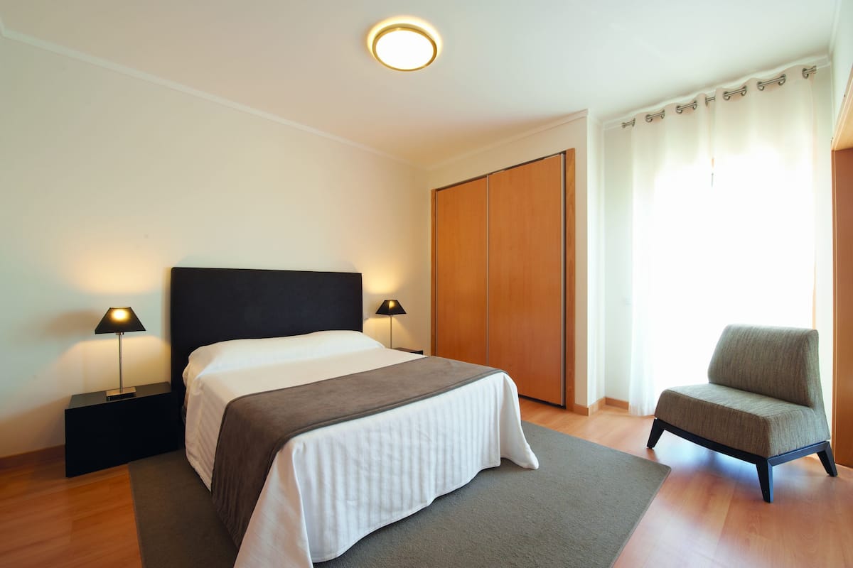 A bedroom with a bed, two bedside tables with lamps, a chair, a wardrobe, and a window with curtains.
