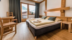 Ein Hotelzimmer mit einem Bett, einem Schreibtisch und einem Stuhl. Es gibt ein Fenster mit Vorhängen, einen Blick auf die Berge und eine Holzbalkendecke.